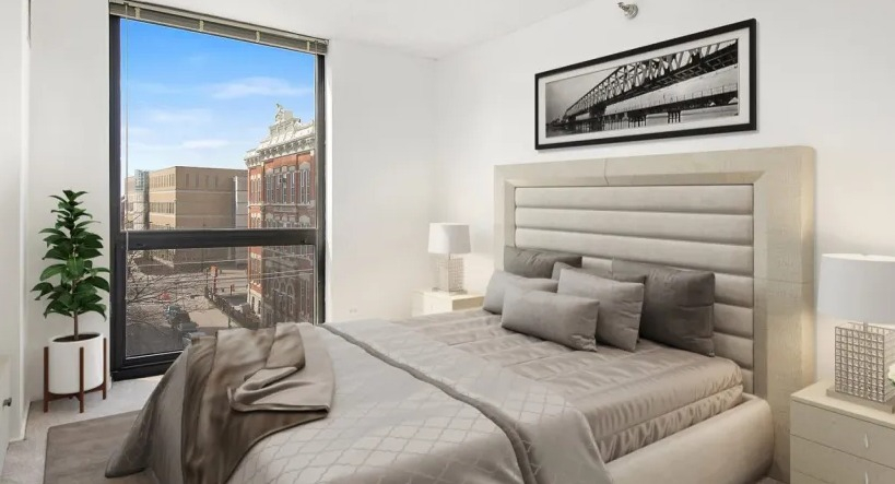 Bright bedroom with an upholstered bed, neutral bedding, nightstands, and a floor-to-ceiling window overlooking the neighborhood at 1000 N LaSalle, Chicago