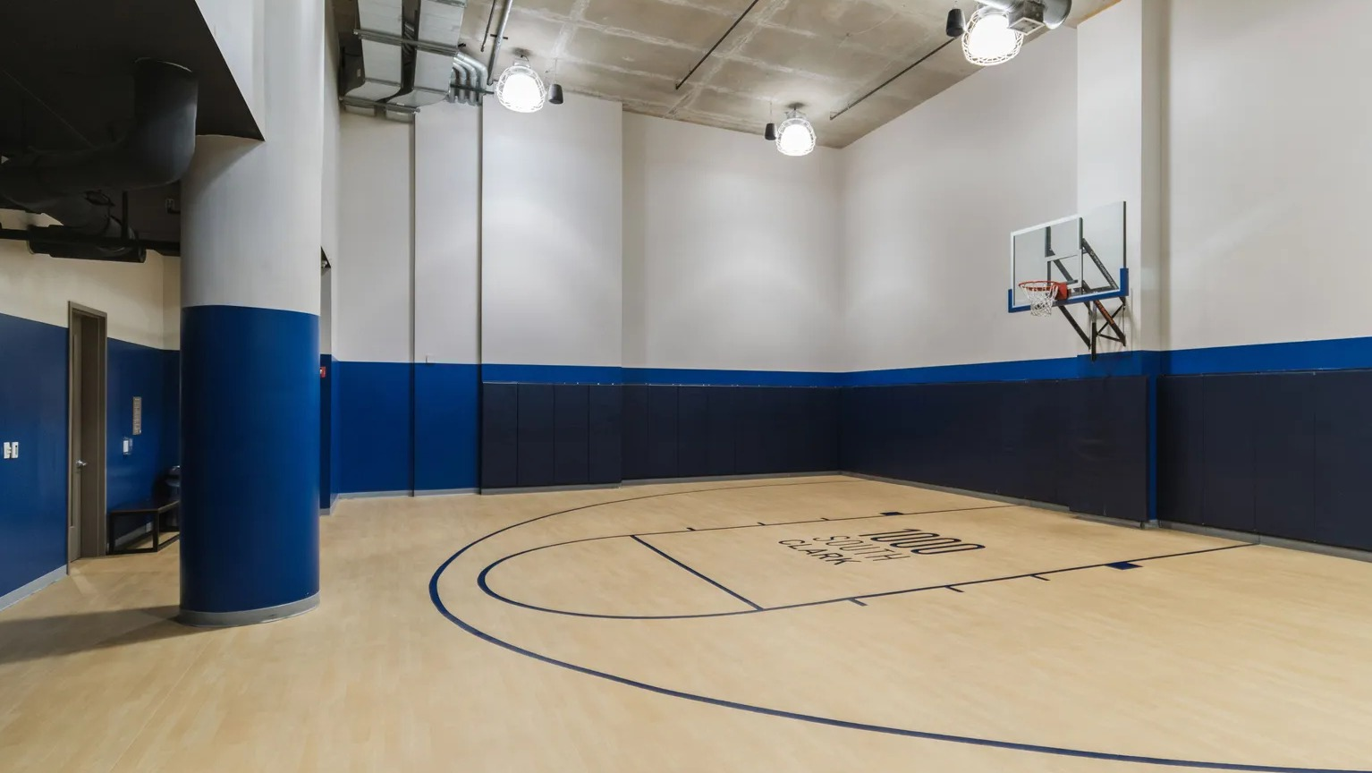 Full-height indoor basketball court with wood-look floor, blue wall padding, and hoop for year-round play at 1000 South Clark apartments in Chicago