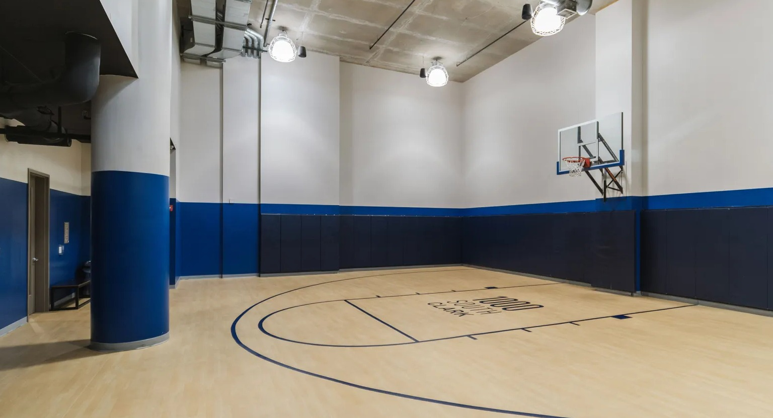 Full-height indoor basketball court with wood-look floor, blue wall padding, and hoop for year-round play at 1000 South Clark apartments in Chicago
