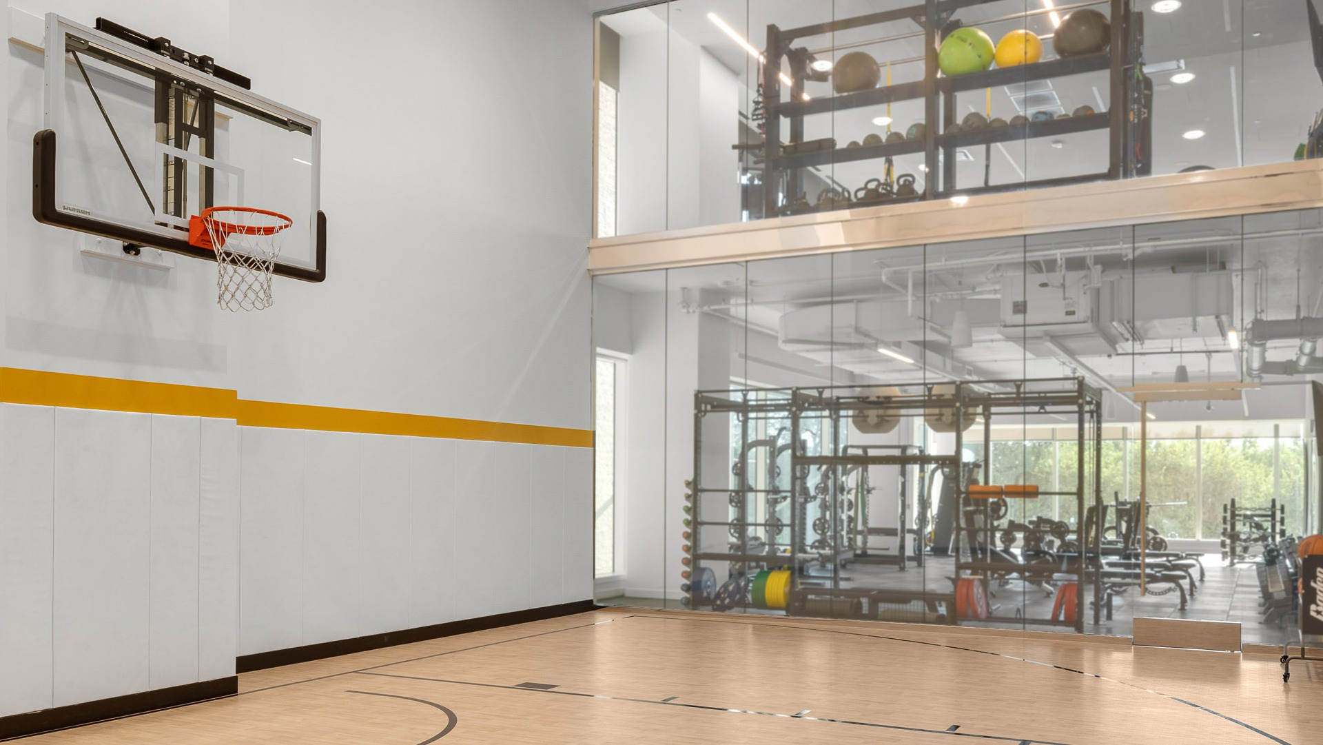 Bright indoor half-court with wall-mounted hoop, maple flooring, and a glass wall overlooking the fitness center at 1000M Apartments in Chicago
