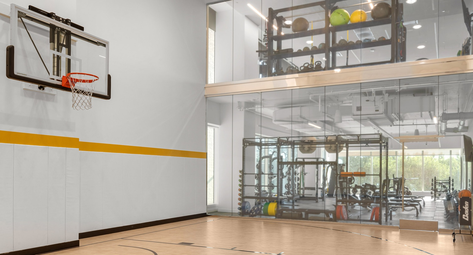 Bright indoor half-court with wall-mounted hoop, maple flooring, and a glass wall overlooking the fitness center at 1000M Apartments in Chicago