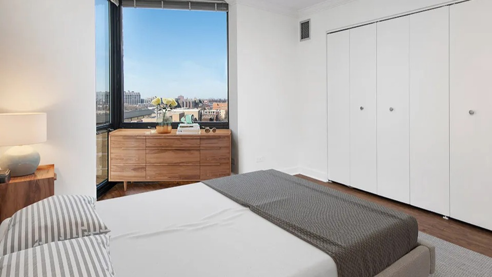 Bedroom with large closet doors, wood dresser, and window overlooking city views at 1120 N LaSalle apartments in Chicago