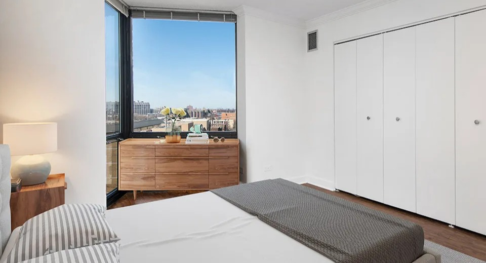 Bedroom with large closet doors, wood dresser, and window overlooking city views at 1120 N LaSalle apartments in Chicago