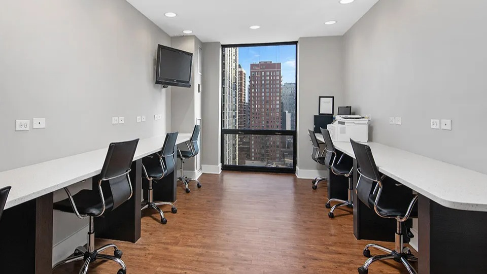 Business center with long workstations, swivel chairs, and a tall window facing city views at 1120 N LaSalle apartments in Chicago