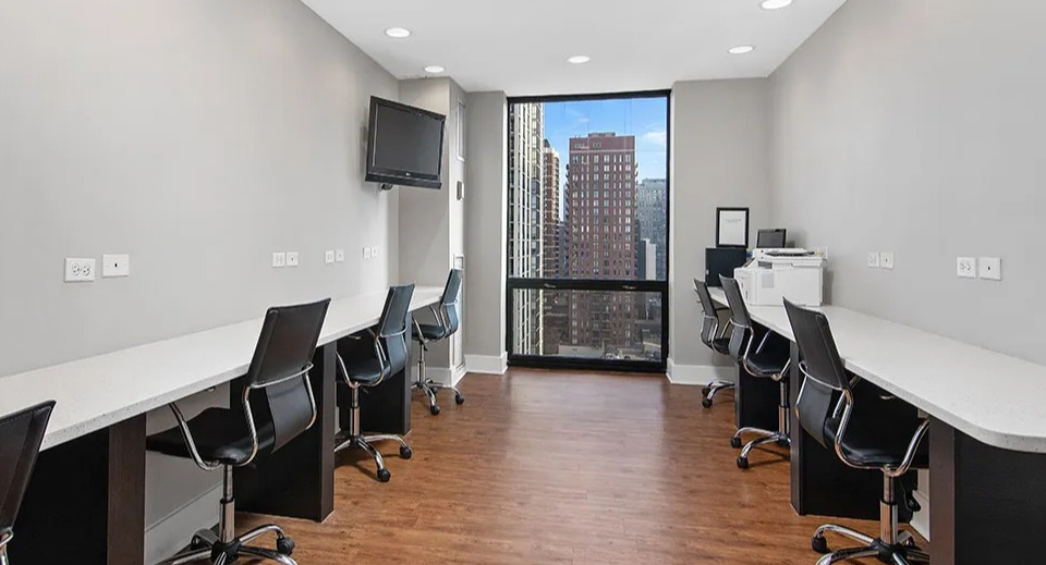 Business center with long workstations, swivel chairs, and a tall window facing city views at 1120 N LaSalle apartments in Chicago