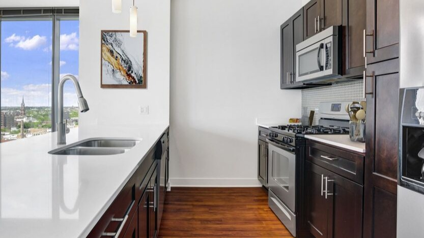 Modern kitchen with long white island, dual sink, dark cabinets, and stainless appliances near tall windows at 1225 Old Town Apartments in Chicago
