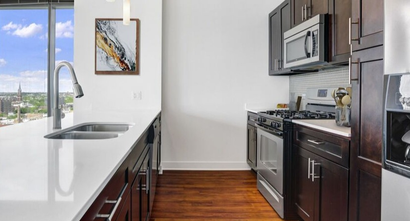 Modern kitchen with long white island, dual sink, dark cabinets, and stainless appliances near tall windows at 1225 Old Town Apartments in Chicago