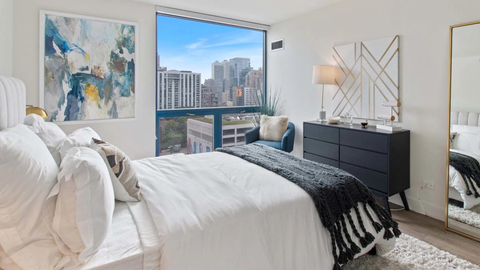 Sunlit bedroom at 1250 LaSalle featuring a full-height mirror, modern dresser, accent chair, and skyline views through an expansive window