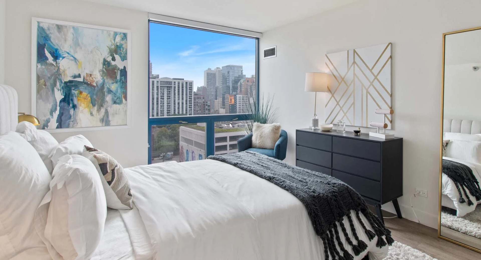 Sunlit bedroom at 1250 LaSalle featuring a full-height mirror, modern dresser, accent chair, and skyline views through an expansive window