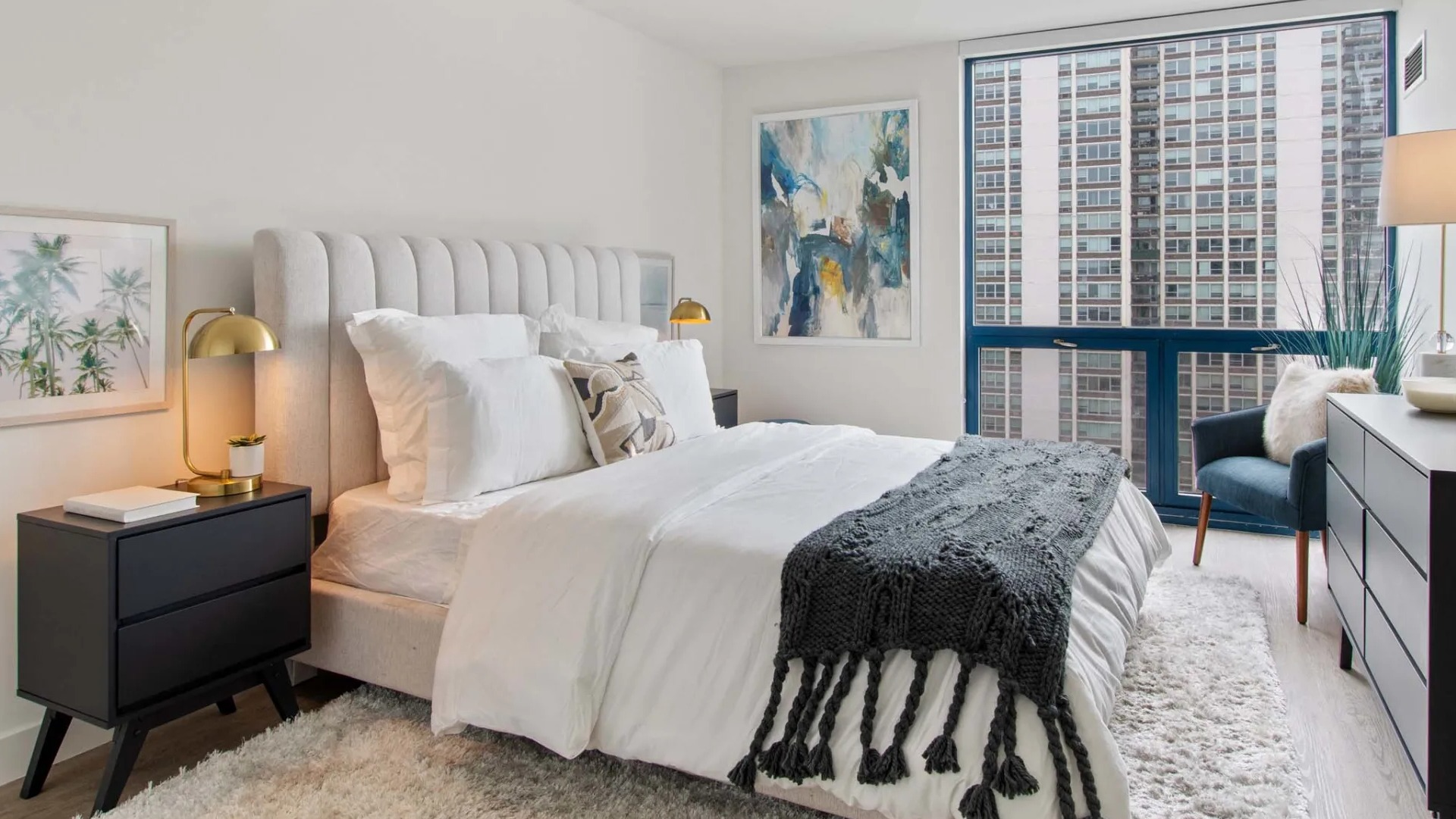 Cozy bedroom at 1250 LaSalle with upholstered headboard, shag rug, black dresser, and a large window framing downtown Chicago skyline views