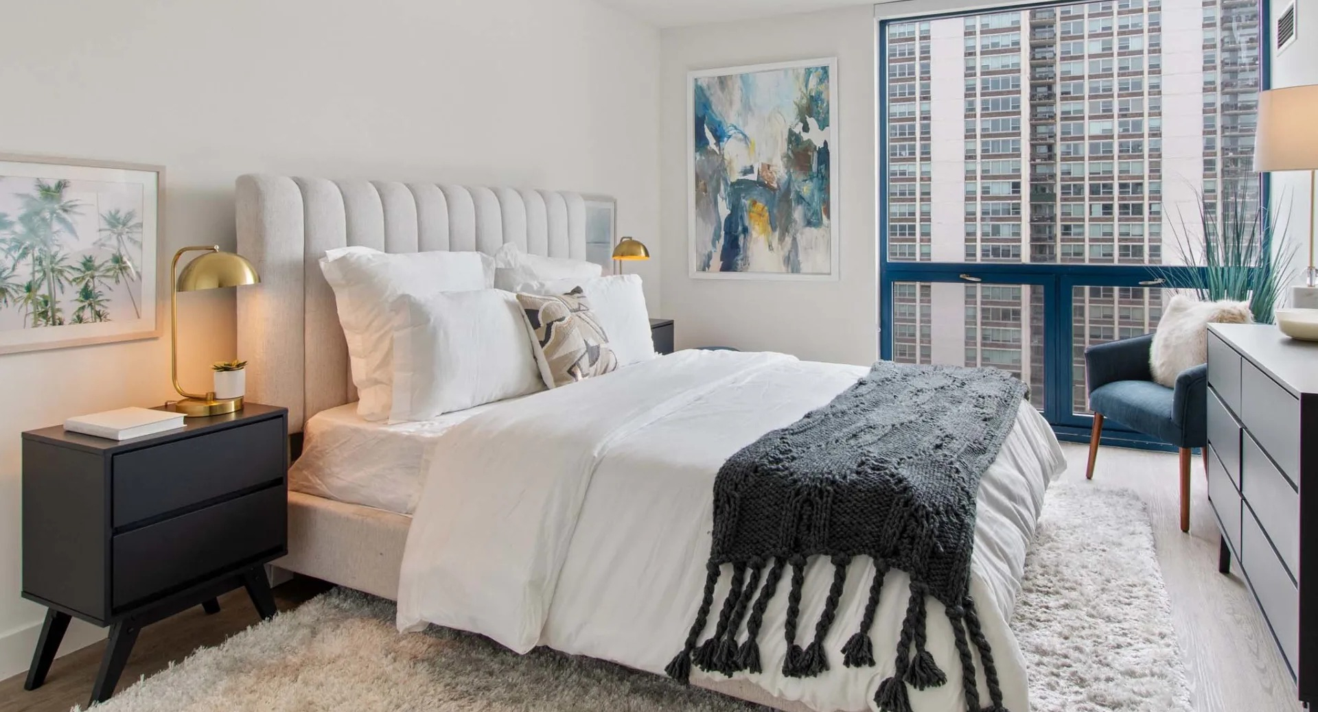 Cozy bedroom at 1250 LaSalle with upholstered headboard, shag rug, black dresser, and a large window framing downtown Chicago skyline views