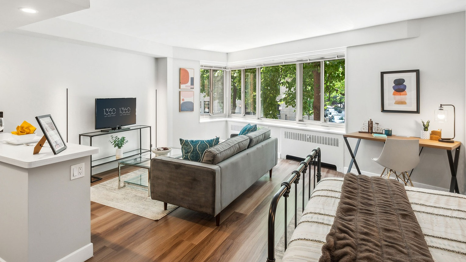 Sunlit studio living area with large corner windows, sofa, TV console, and work desk at 1350–1360 Lake Shore Drive apartments in Chicago