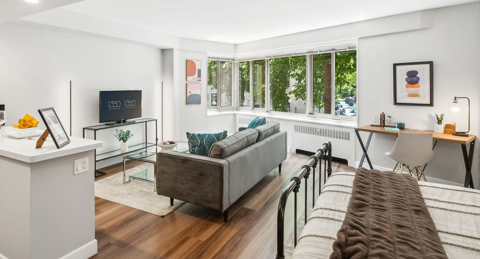 Sunlit studio living area with large corner windows, sofa, TV console, and work desk at 1350–1360 Lake Shore Drive apartments in Chicago