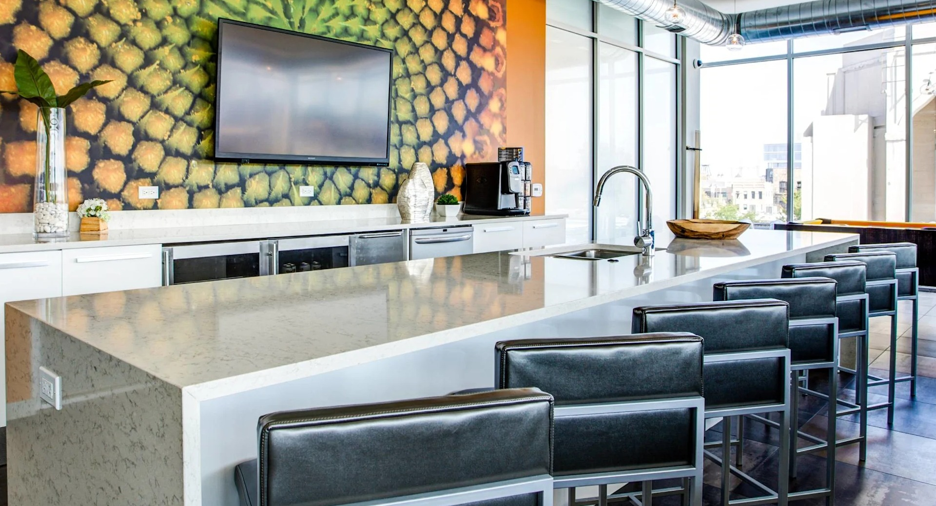 Stylish clubroom kitchen at 13thirteen Randolph Street Lofts with long quartz island, bar seating, TV, and bold accent wall overlooking Chicago views