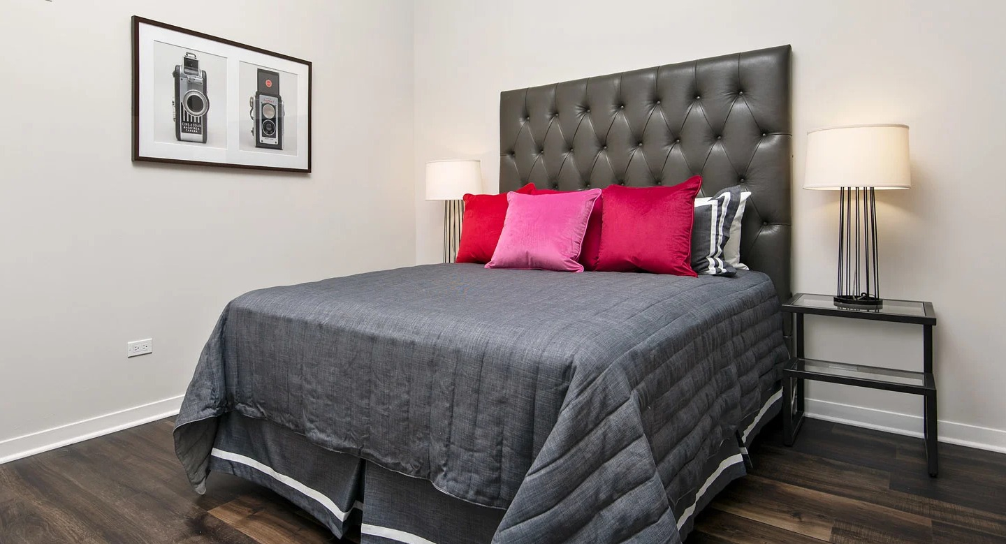 Stylish bedroom at 13thirteen Randolph Street Lofts featuring tufted headboard, dark bedding, bright accent pillows, and wood-look floors in Chicago