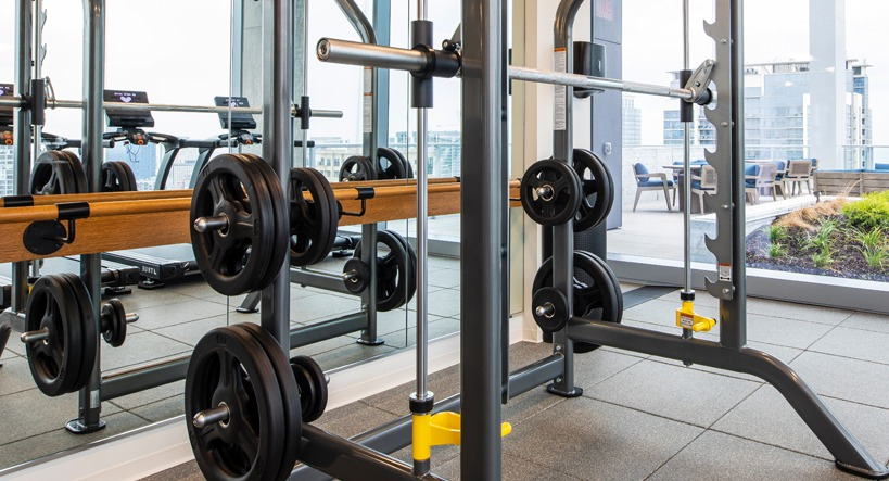 Fitness center weight room with Smith machine, mirrored wall, and cardio equipment beside floor-to-ceiling windows at 1400 Wabash apartments in Chicago