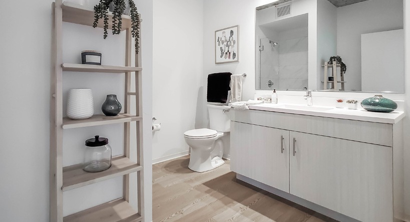Bright bathroom with double vanity, quartz counters, open wood shelving, and a sleek glass shower at 1400 Wabash apartments in Chicago