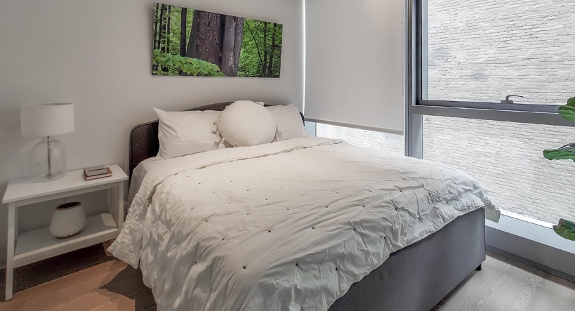 Bright bedroom with queen bed, crisp white bedding, roller shades, and a large window bringing natural light at 1400 Wabash in Chicago