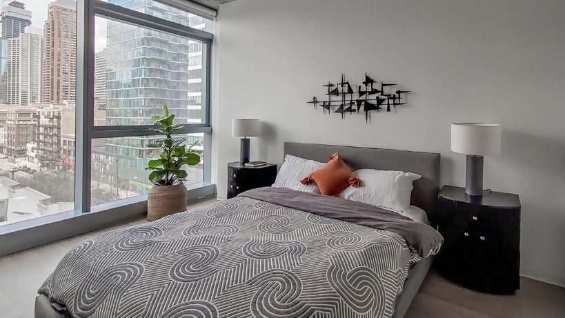 Corner bedroom with floor-to-ceiling windows, skyline views, and matching lamps flanking a low bed at 1400 Wabash apartments in Chicago