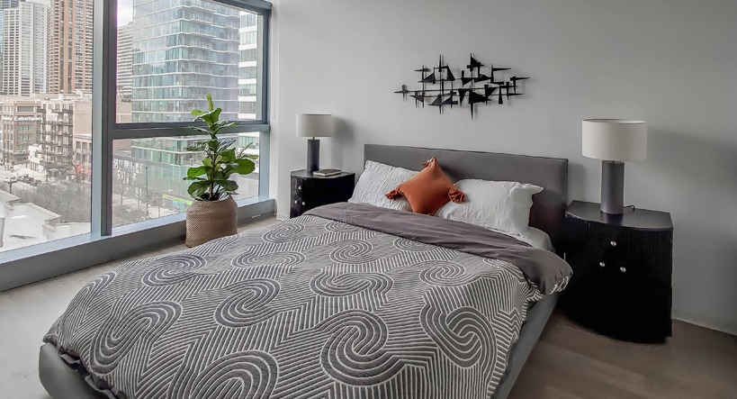 Corner bedroom with floor-to-ceiling windows, skyline views, and matching lamps flanking a low bed at 1400 Wabash apartments in Chicago