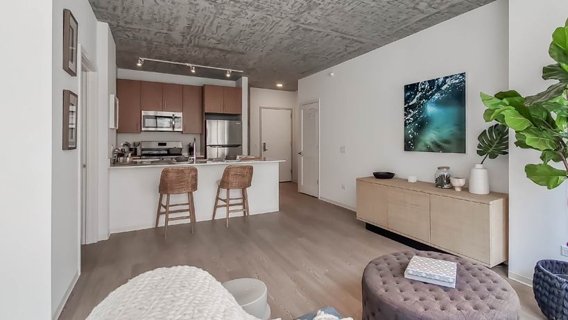 Spacious living room with large plant, media console, soft seating, and track lighting near the kitchen at 1400 Wabash apartments in Chicago