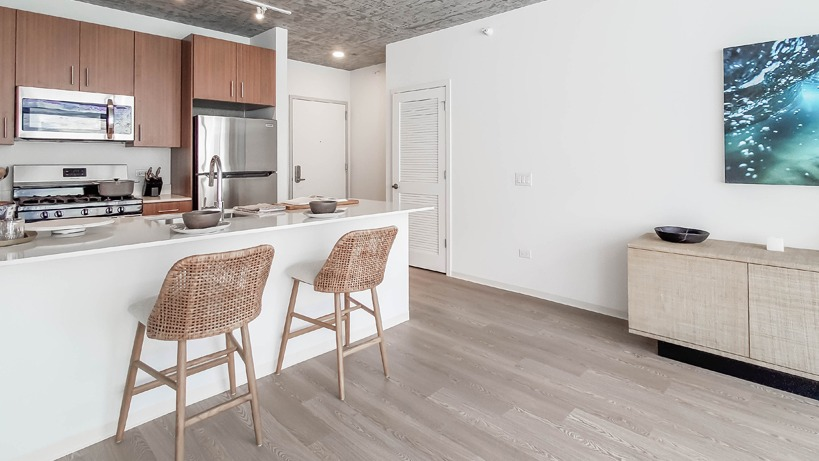 Open living area facing the kitchen island with woven stools, stainless appliances, and bright finishes at 1400 Wabash apartments in Chicago