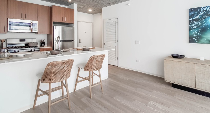 Open living area facing the kitchen island with woven stools, stainless appliances, and bright finishes at 1400 Wabash apartments in Chicago