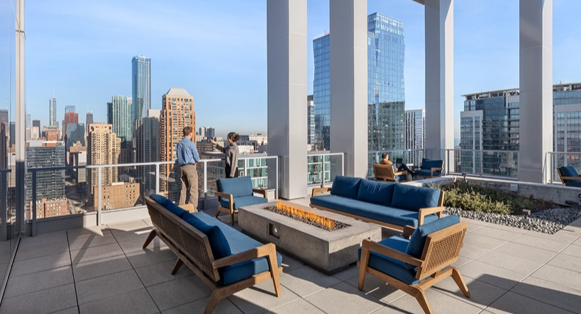 Rooftop deck lounge with blue cushioned sofas around a modern fire pit, tall frame columns, and sweeping skyline views at 1400 Wabash in Chicago