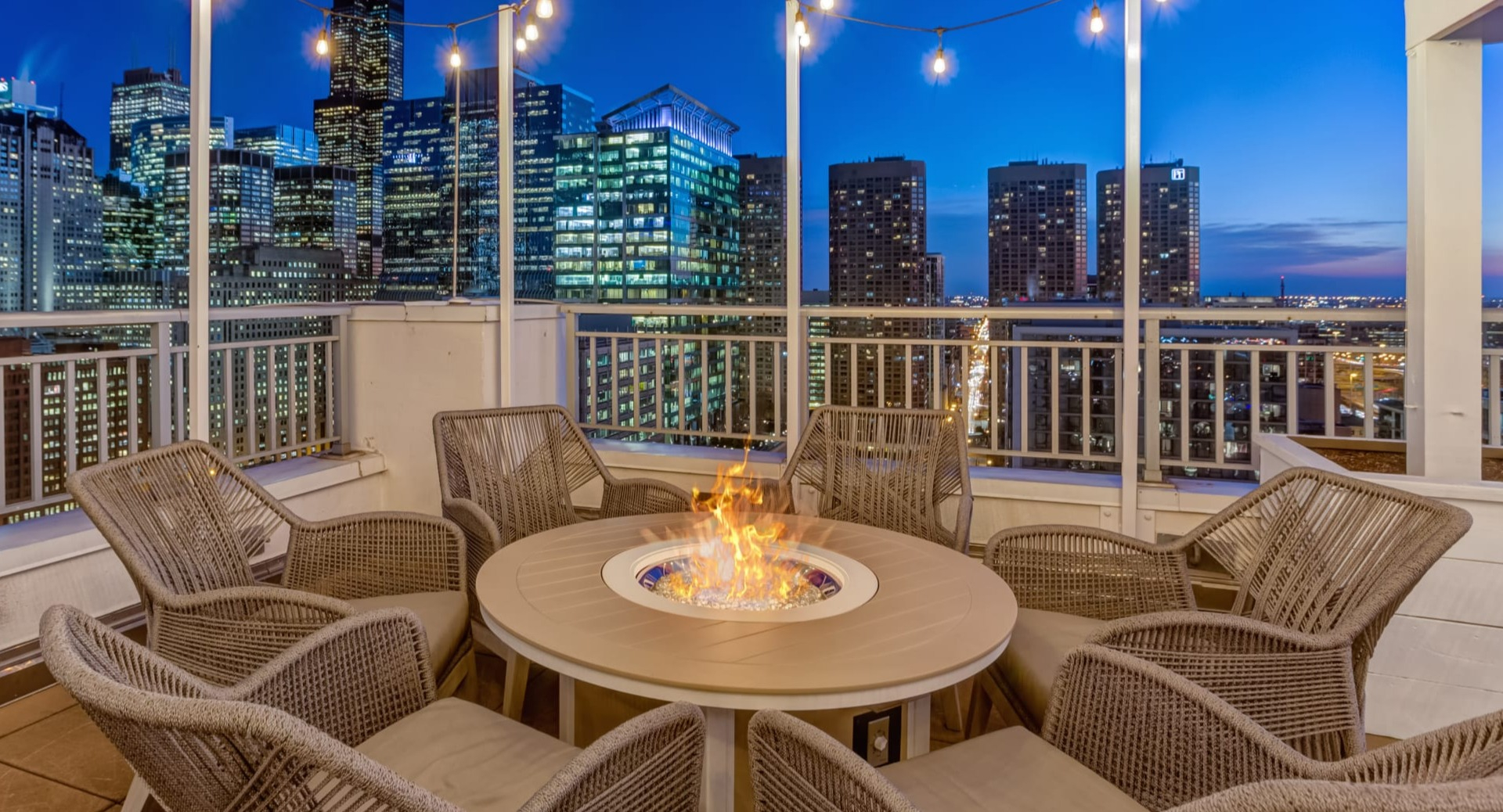 Rooftop terrace with round fire pit, wicker lounge chairs and string lights set against downtown skyline at 180 North Jefferson apartments in Chicago