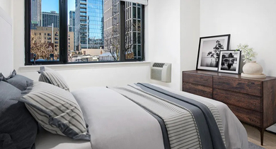 Bright bedroom with city views through wide windows, a modern wood dresser, and layered bedding at 20 E Scott apartments in Chicago