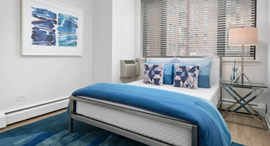 Cozy bedroom with blue bedding, geometric pillows, large window with blinds, and a metal frame bed at 20 E Scott apartments in Chicago