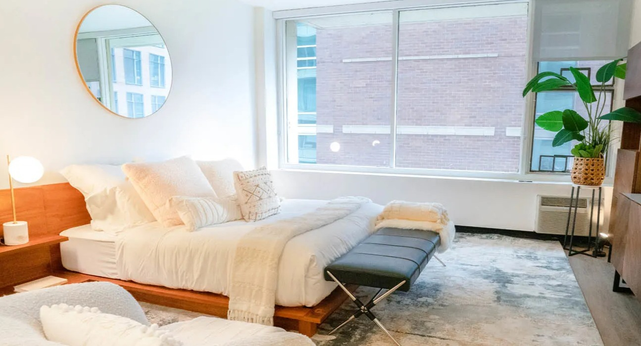 Spacious studio bedroom with large window, wood platform bed, bench seating, abstract rug, and media console at 21 E Chestnut in Chicago