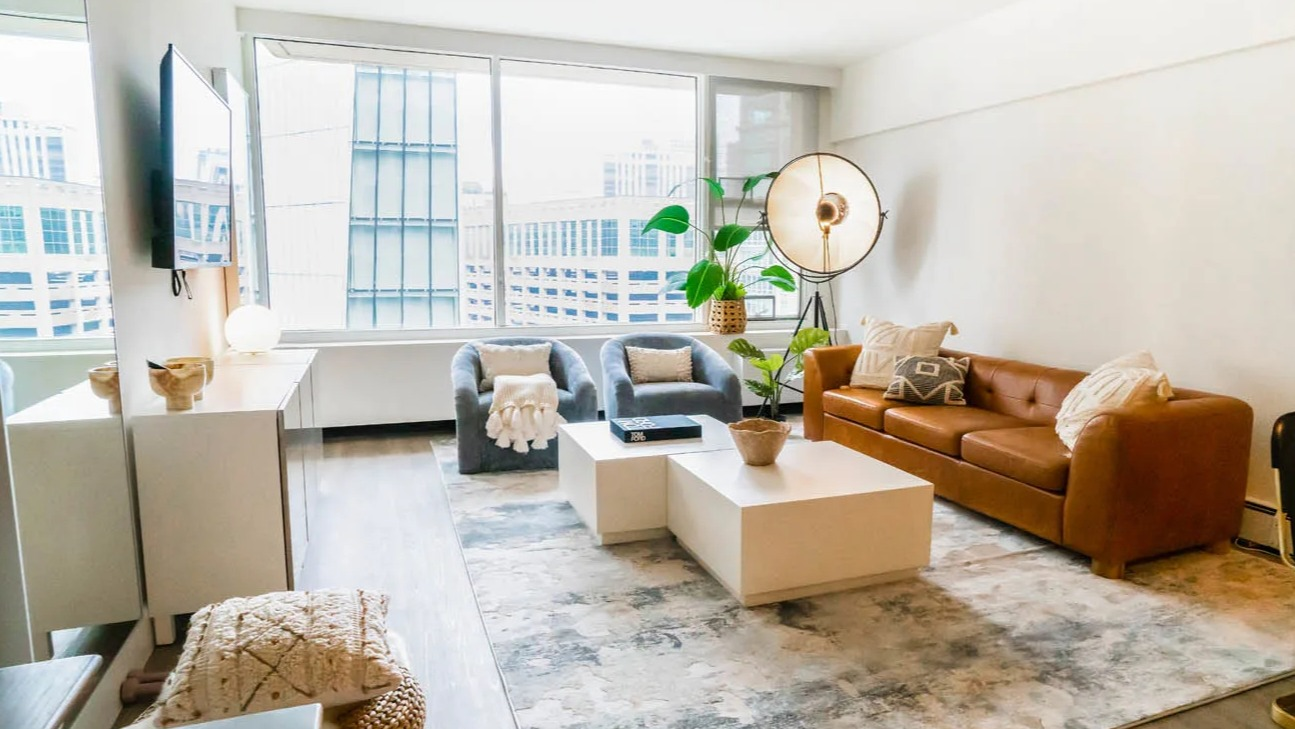 Sunlit living room with floor-to-ceiling windows, caramel sofa, accent chairs, and textured rug at 21 E Chestnut apartments in Chicago