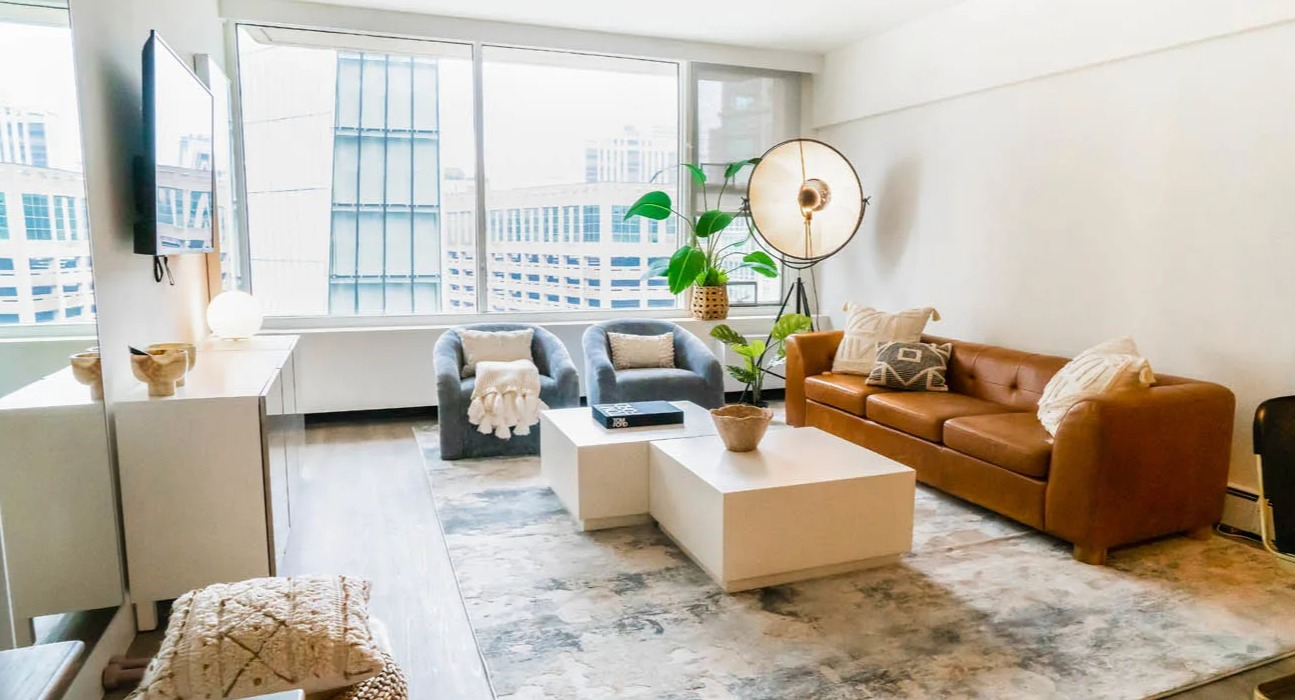 Sunlit living room with floor-to-ceiling windows, caramel sofa, accent chairs, and textured rug at 21 E Chestnut apartments in Chicago