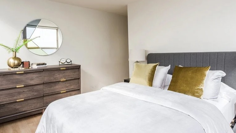 Cozy bedroom with a gray upholstered headboard, crisp white bedding, and gold accent pillows at 2101 South Michigan Apartments in Chicago, styled with a dresser and mirror