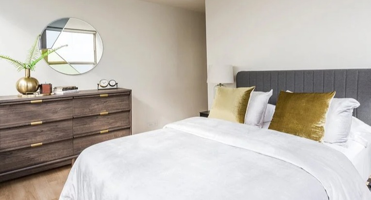 Cozy bedroom with a gray upholstered headboard, crisp white bedding, and gold accent pillows at 2101 South Michigan Apartments in Chicago, styled with a dresser and mirror