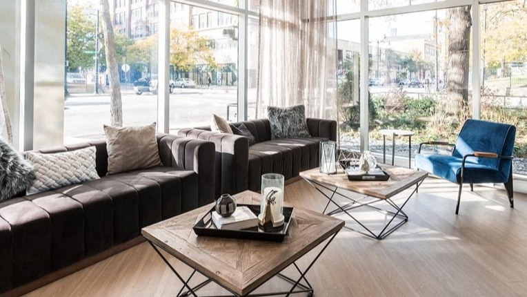Sunlit resident lounge with a long brown sectional, modern chandelier, and geometric coffee tables by floor-to-ceiling windows at 2101 South Michigan Apartments in Chicago