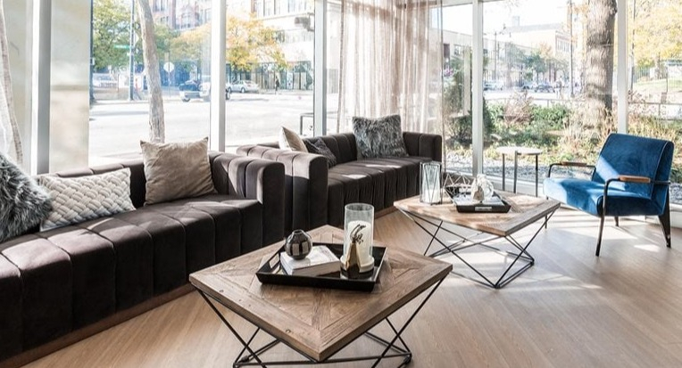 Sunlit resident lounge with a long brown sectional, modern chandelier, and geometric coffee tables by floor-to-ceiling windows at 2101 South Michigan Apartments in Chicago
