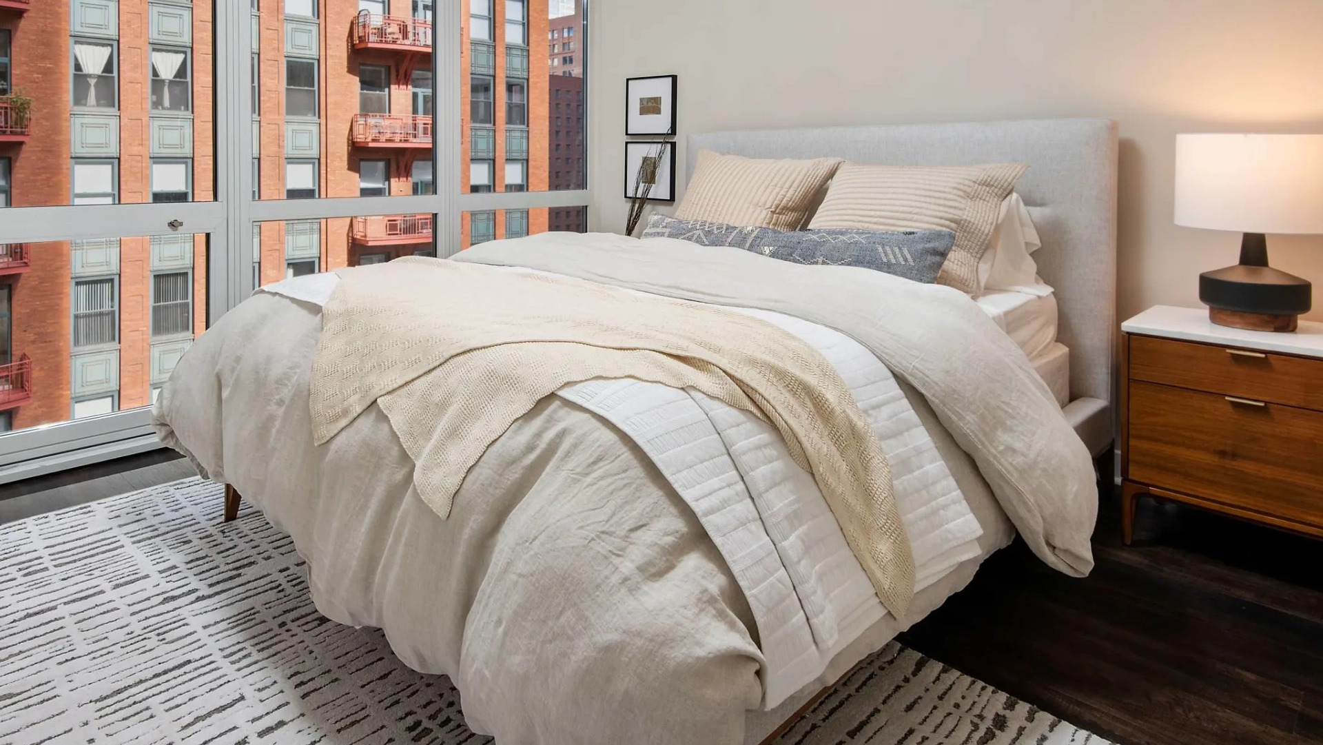 Cozy bedroom at 215 West Chicago with upholstered bed, nightstand lamp, and floor-to-ceiling windows overlooking nearby brick buildings and balconies