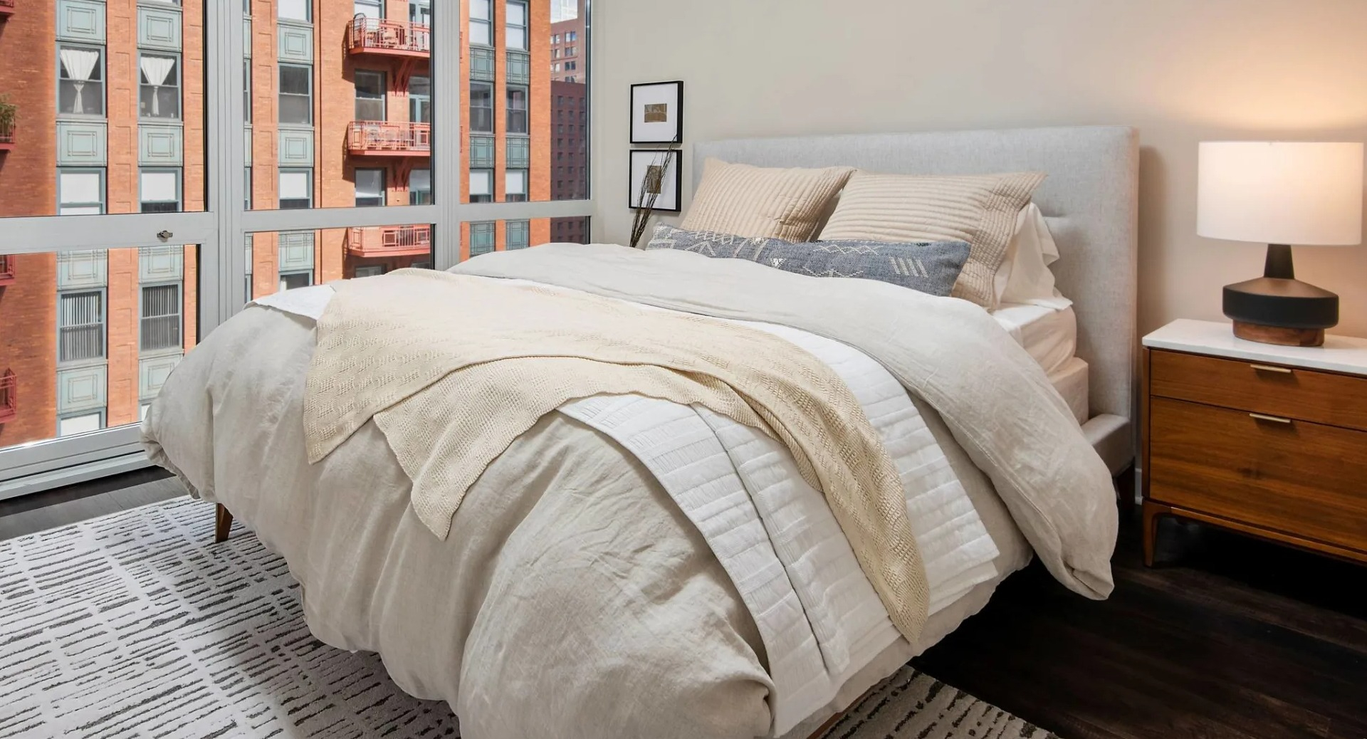 Cozy bedroom at 215 West Chicago with upholstered bed, nightstand lamp, and floor-to-ceiling windows overlooking nearby brick buildings and balconies