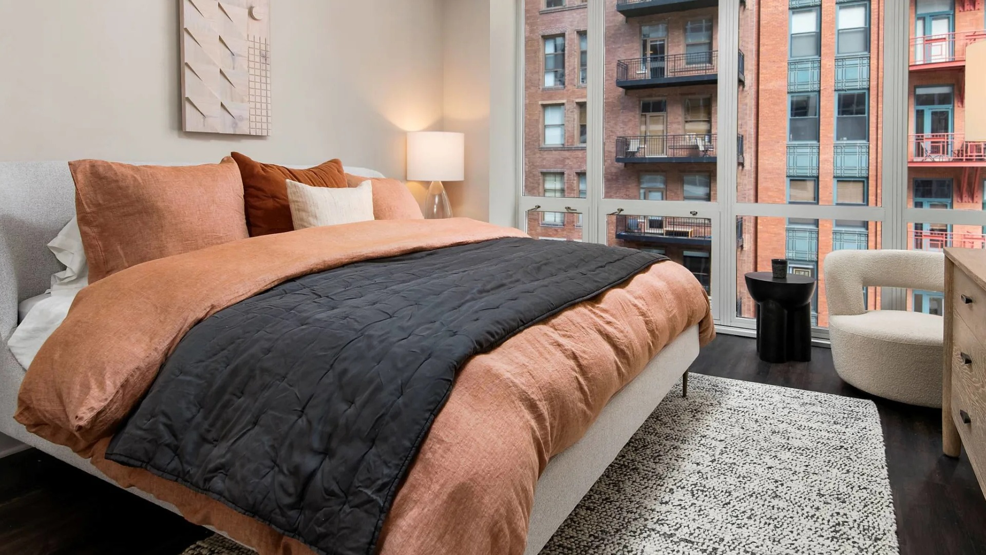 Spacious primary bedroom at 215 West in Chicago showing a large bed with warm linens, dresser and chair, and wall-to-wall windows with city views