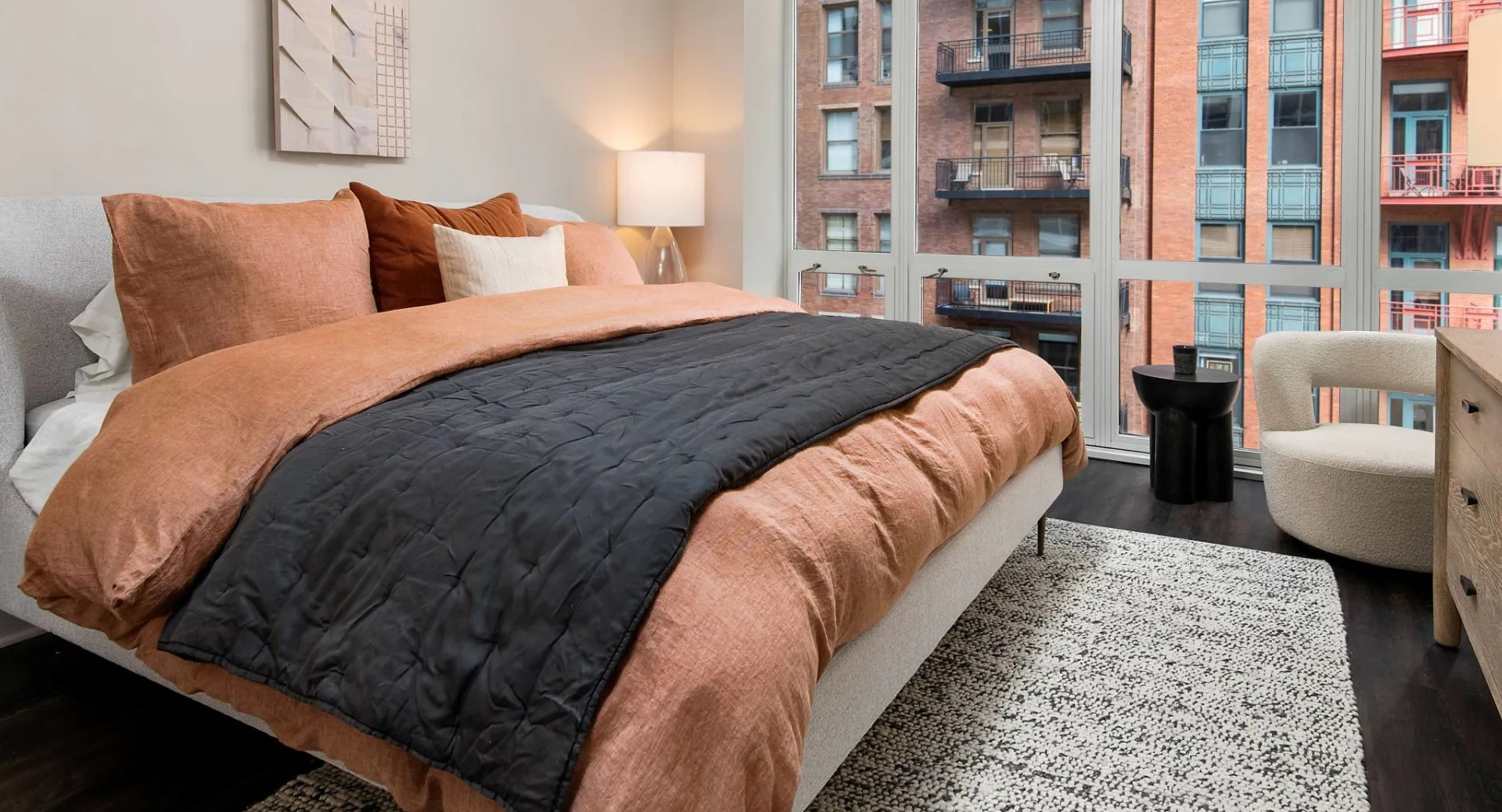 Spacious primary bedroom at 215 West in Chicago showing a large bed with warm linens, dresser and chair, and wall-to-wall windows with city views