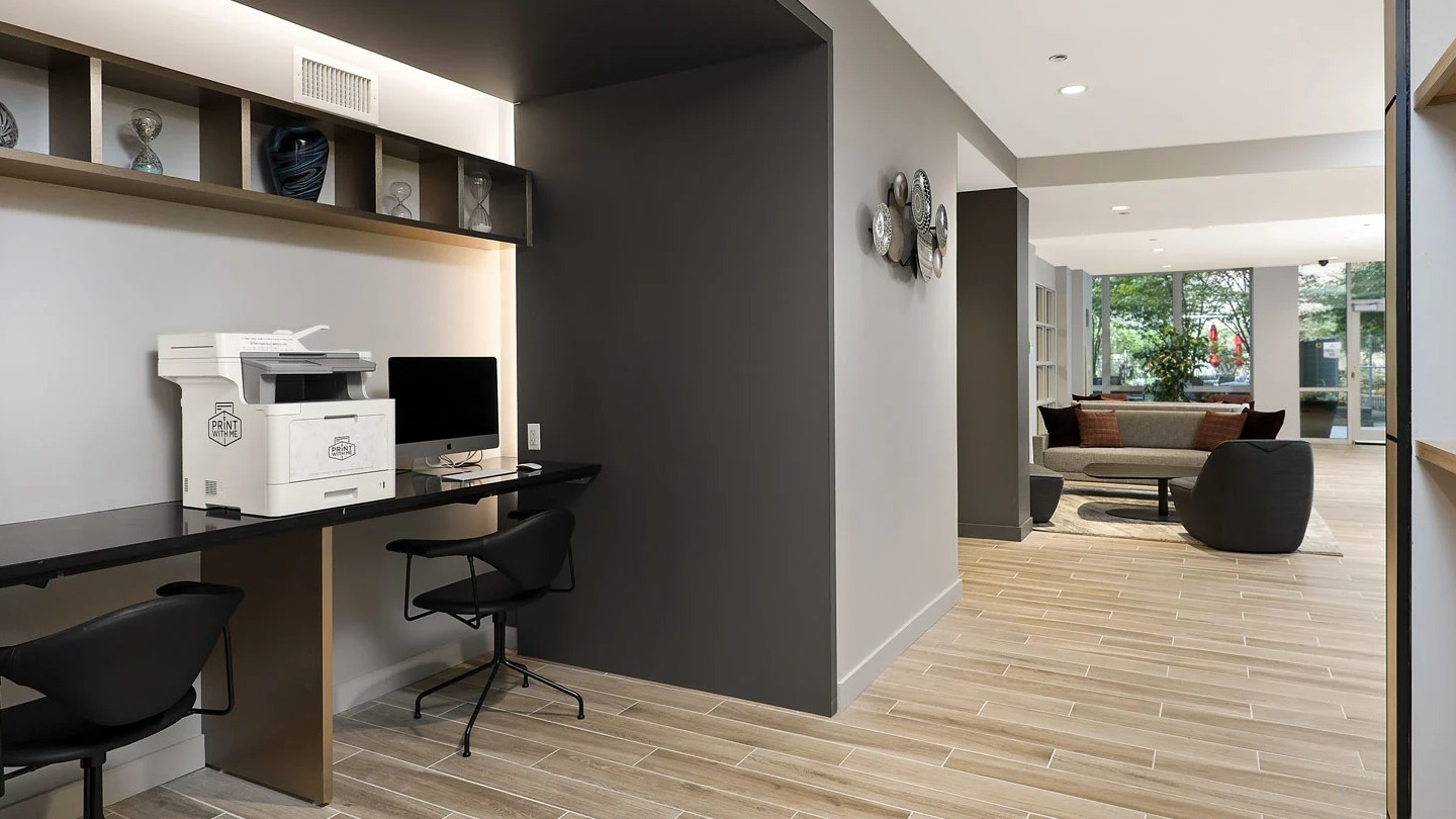 Business center at 215 West in Chicago with computer workstations, printer, long desk, shelving, and bright lounge access for remote work