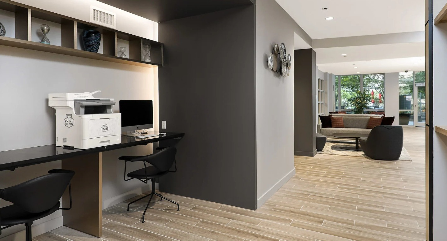 Business center at 215 West in Chicago with computer workstations, printer, long desk, shelving, and bright lounge access for remote work