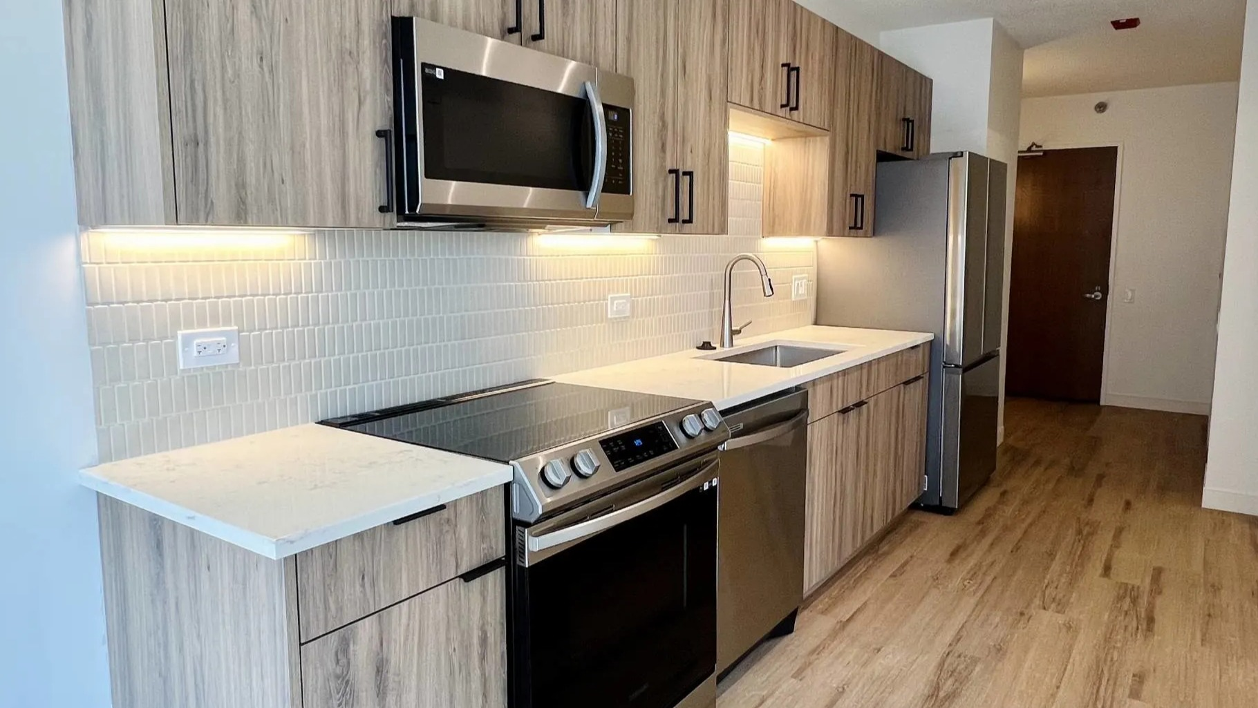 Renovated studio kitchen at 215 West Chicago with wood-grain cabinets, quartz counters, tile backsplash, stainless appliances, and under-cabinet lights