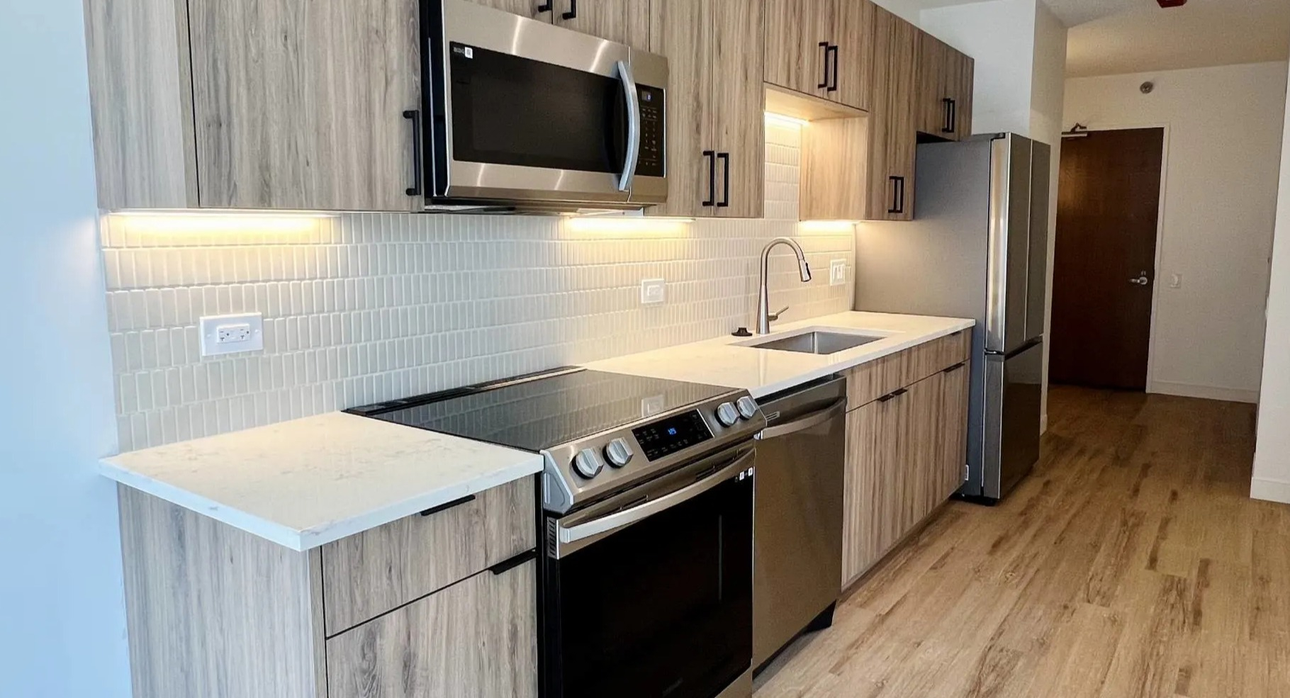 Renovated studio kitchen at 215 West Chicago with wood-grain cabinets, quartz counters, tile backsplash, stainless appliances, and under-cabinet lights