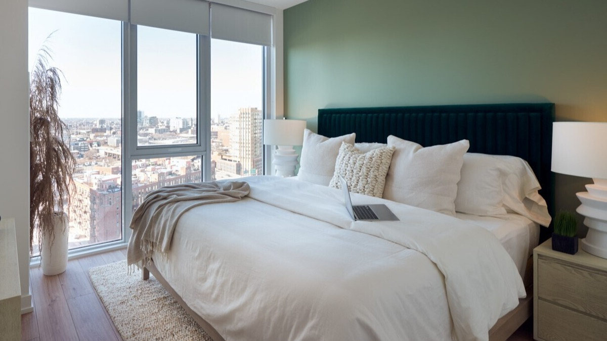 Bright bedroom with soft green accent wall, velvet headboard, large corner windows, and neutral bedding at 369 Grand apartments in Chicago