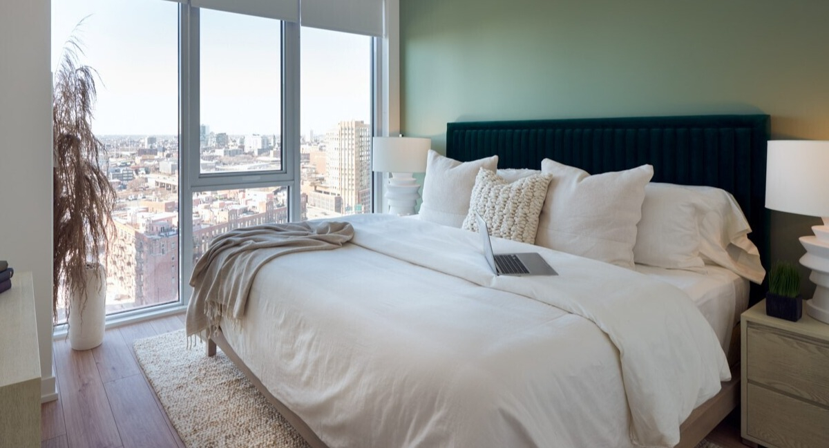 Bright bedroom with soft green accent wall, velvet headboard, large corner windows, and neutral bedding at 369 Grand apartments in Chicago