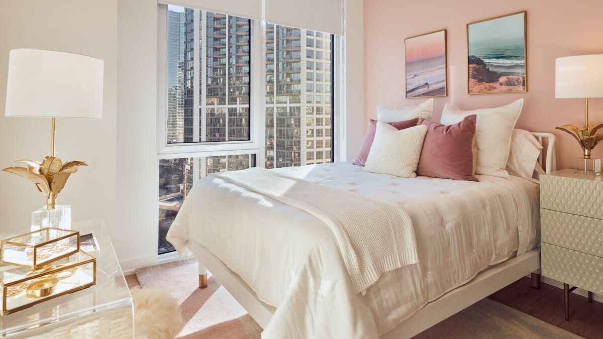 Cozy bedroom with blush accent wall, gold decor, soft white bedding, and large windows with downtown views at 369 Grand apartments in Chicago