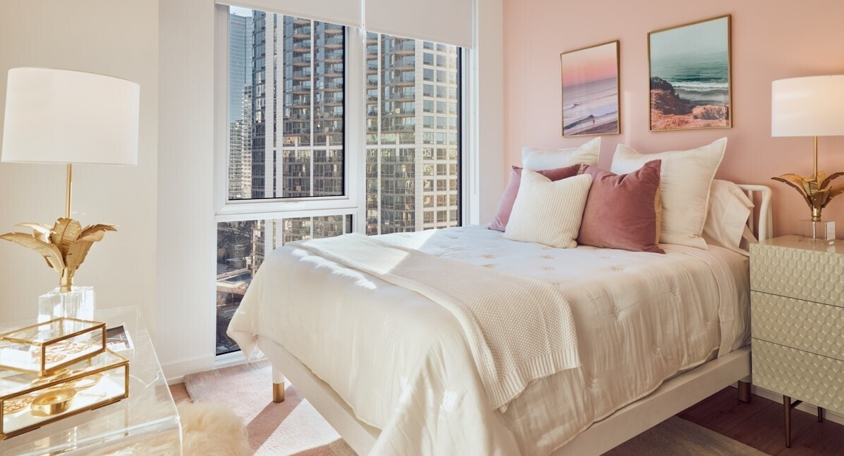 Cozy bedroom with blush accent wall, gold decor, soft white bedding, and large windows with downtown views at 369 Grand apartments in Chicago